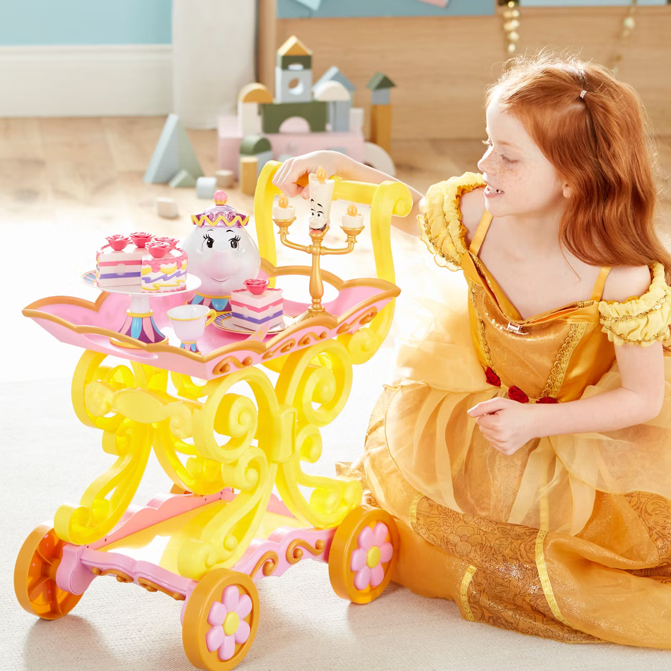 Child in a yellow dress playing with a toy tea set and cake in a bright room.